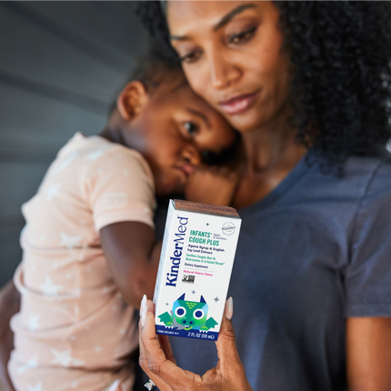 Image of a woman holding her child, and reading the back of the KinderMed Infants Cough Plus packaging.